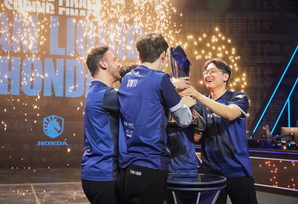 Esports-team in blauwe jerseys viert overwinning op het podium met trofee en confetti tijdens een levendig kampioenschapsevenement.