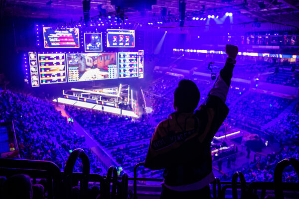 Fan juicht voor een esports-wedstrijd in een grote arena, met een overzicht van het verlichte podium en schermen die de score tonen.