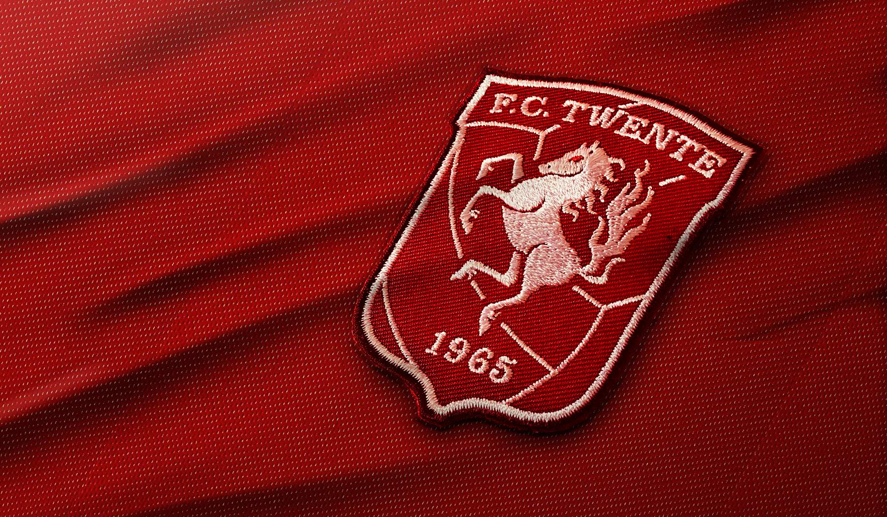 Het logo van FC Twente, een Nederlandse voetbalclub uit Enschede, geborduurd op een rode stof. Het logo toont een steigerend paard, een symbool van kracht en vastberadenheid, met het jaartal 1965 onder het paard. Dit jaar markeert de oprichting van de club.