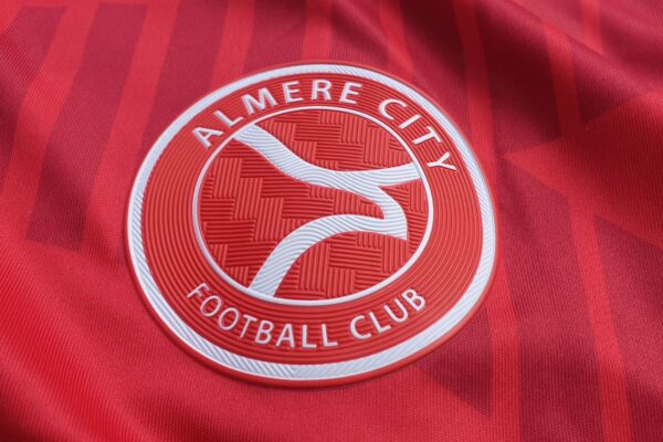 Het beeld toont het logo van Almere City Football Club op hun rode tenue, met een strak en cirkelvormig ontwerp. Het embleem benadrukt de naam van de club met vloeiende lijnen die de ambitie en de band met de stad Almere symboliseren.