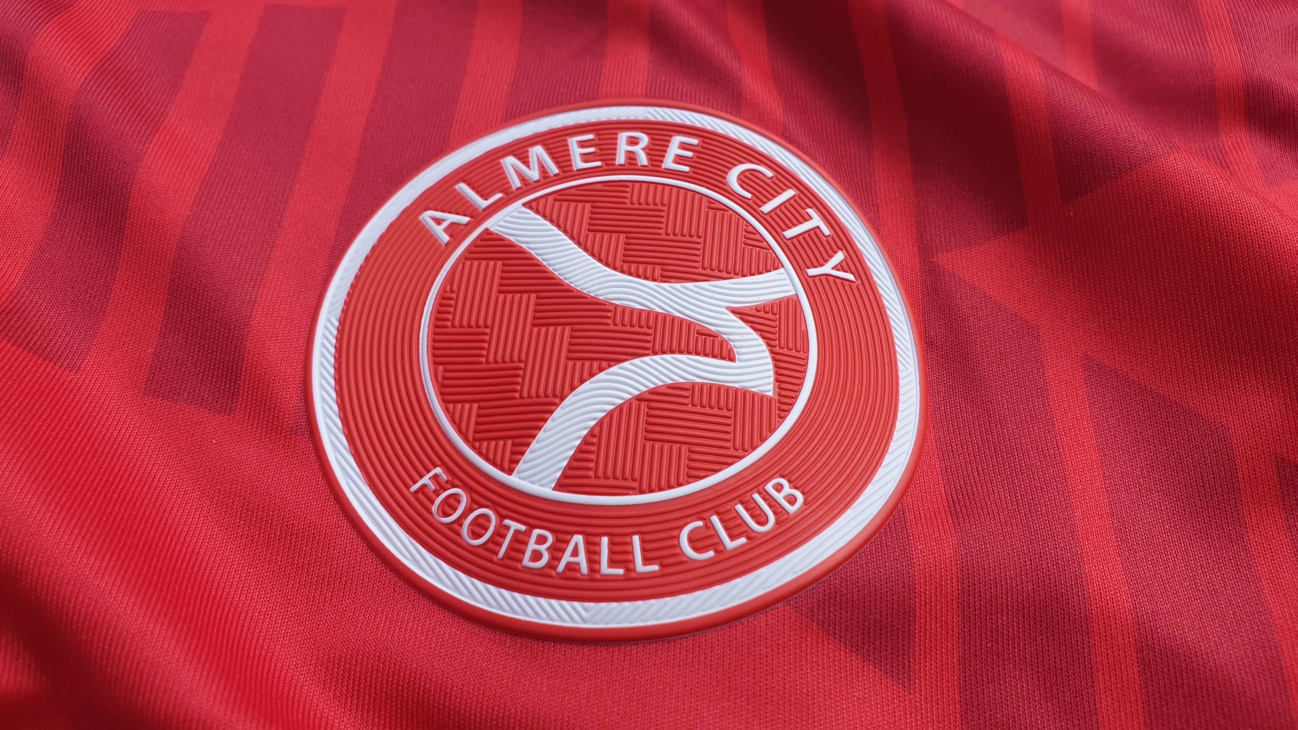 Het beeld toont het logo van Almere City Football Club op hun rode tenue, met een strak en cirkelvormig ontwerp. Het embleem benadrukt de naam van de club met vloeiende lijnen die de ambitie en de band met de stad Almere symboliseren.