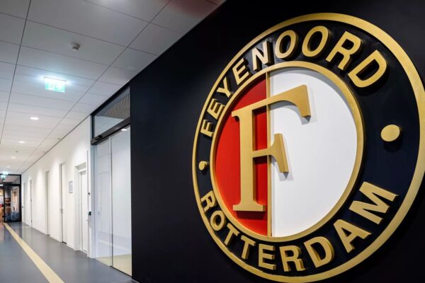 Het logo van Feyenoord Rotterdam aan de muur van het stadion, met de iconische gouden "F" in het midden, omgeven door de clubkleuren rood, wit en zwart.