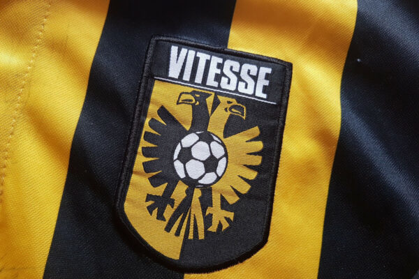 Op het embleem staat de clubnaam “Vitesse” in vette letters boven een gestileerde zwarte adelaar die een voetbal vasthoudt, tegen de traditionele geel met zwart gestreepte achtergrond.